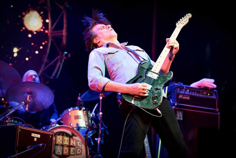 Guitarist in grey shirt performing with green electric guitar under purple stage lighting, with drummer visible in background.