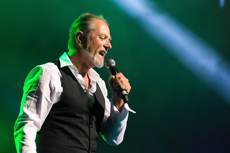 Performer in black vest and white shirt singing into microphone under green stage lighting.