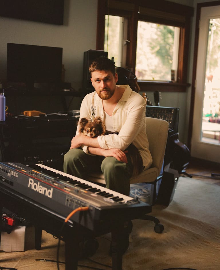 Man seated at a Roland keyboard in a sunlit studio space with windows and recording equipment visible.