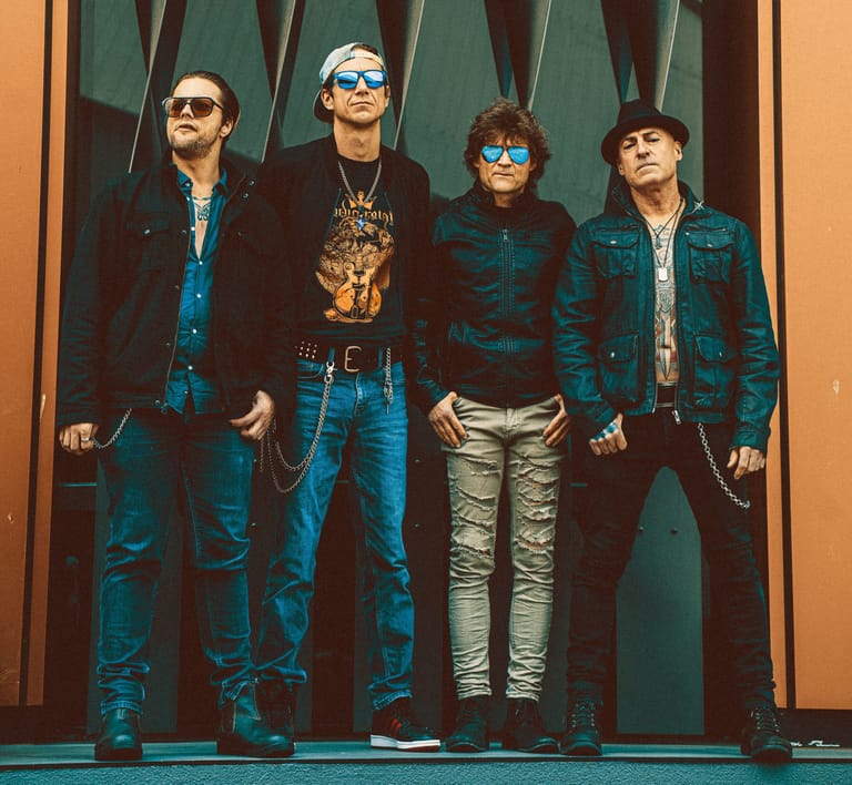 Four band members in denim and sunglasses stand against a green and orange striped backdrop.