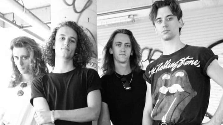 Four band members pose together in black and white, wearing casual dark clothing against an industrial backdrop.