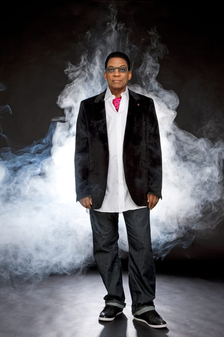 Man in black blazer and pink tie standing amid swirling white smoke against dark background.