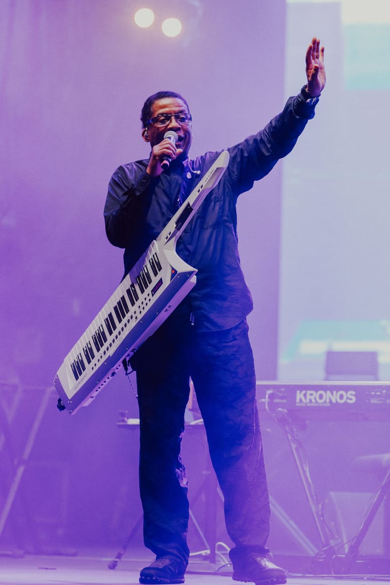 Musician performs with keyboard and microphone on stage beneath purple lighting.