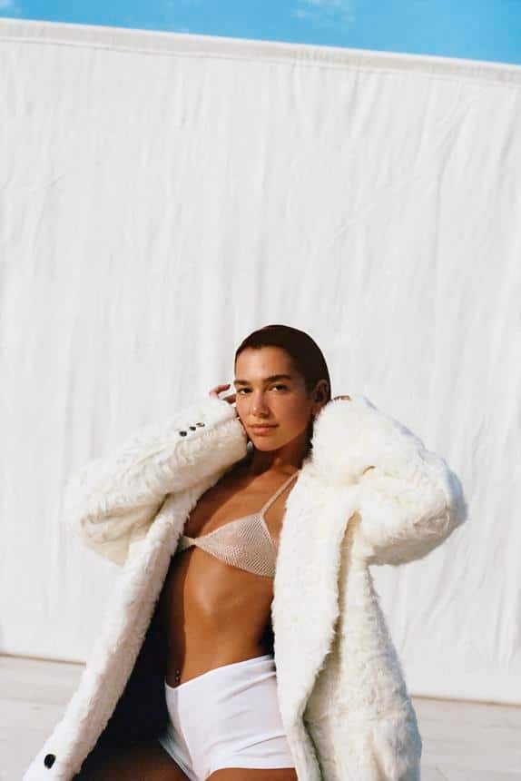 Person in white bikini and cream textured coat posing against white textured wall and blue sky backdrop.