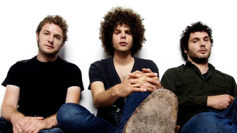 Three men with curly hair sit together against a white background in casual black shirts and jeans.