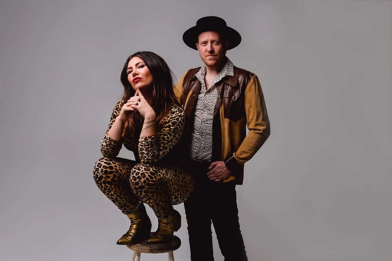 Woman in leopard-print outfit seated on stool beside man in brown leather jacket and black hat against grey background.