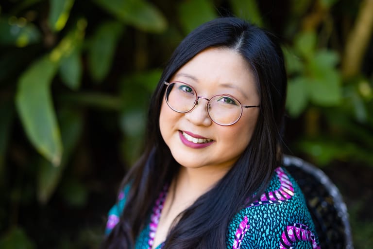 Young woman with long dark hair and round glasses smiling at camera against blurred green foliage.