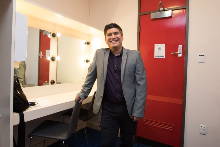 Man in grey blazer and dark shirt standing in a modern dressing room with illuminated mirror and red accent wall.