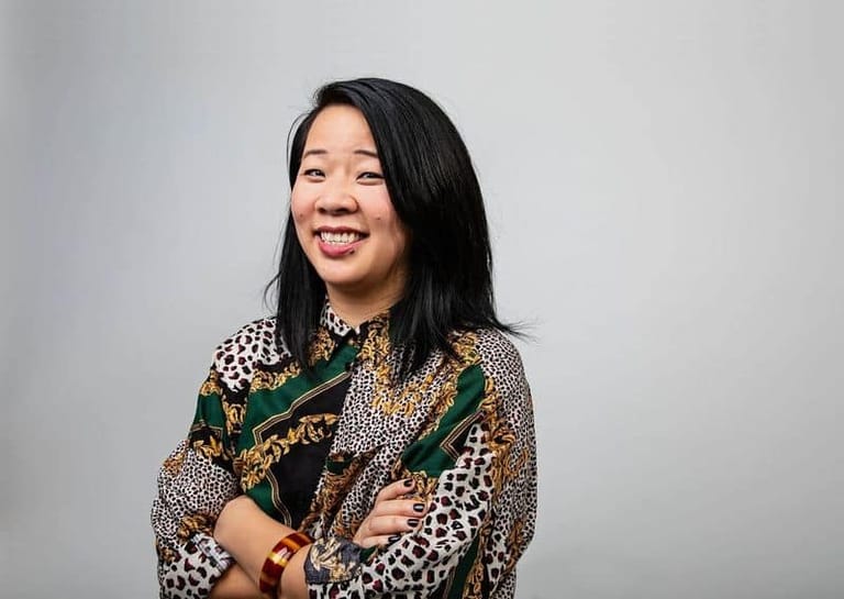 Woman in a patterned shirt with green and gold accents smiling against a neutral grey background.