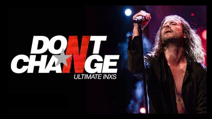 Long-haired male performer in black shirt raises microphone aloft on stage under blue spotlight.