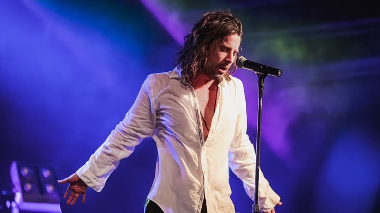 Musician in white shirt singing at microphone beneath blue and purple stage lighting.