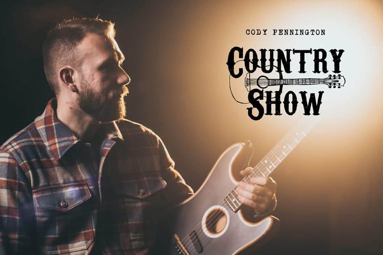 Bearded musician in plaid shirt holding acoustic guitar against warm stage lighting.