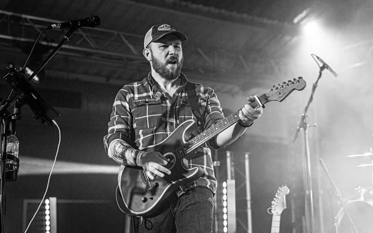 Musician in checked shirt and cap playing electric guitar on stage under bright spotlights.