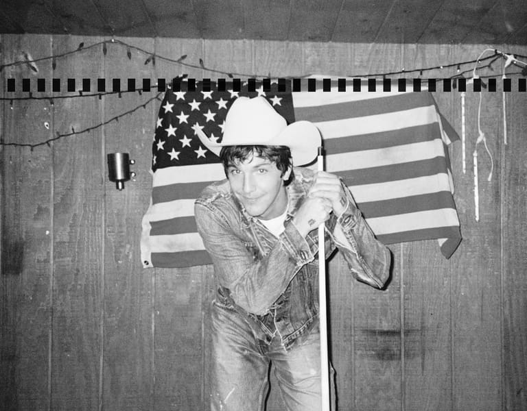 Performer in white cowboy hat and sequined jacket singing at microphone against American flag backdrop.