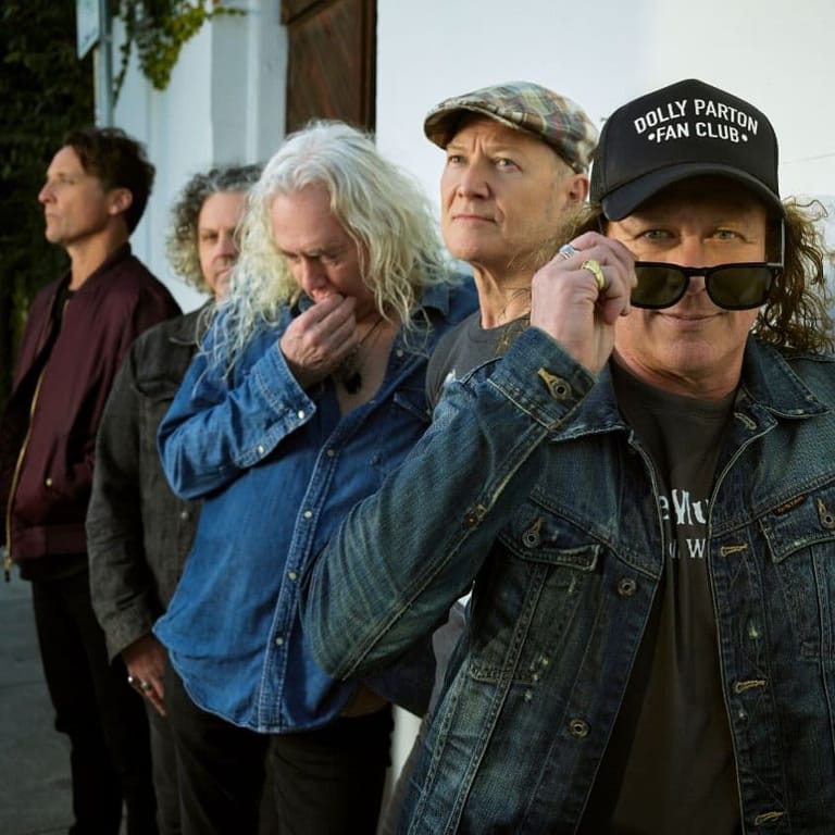 Group of five musicians wearing denim and casual wear stand together outdoors against a white building.