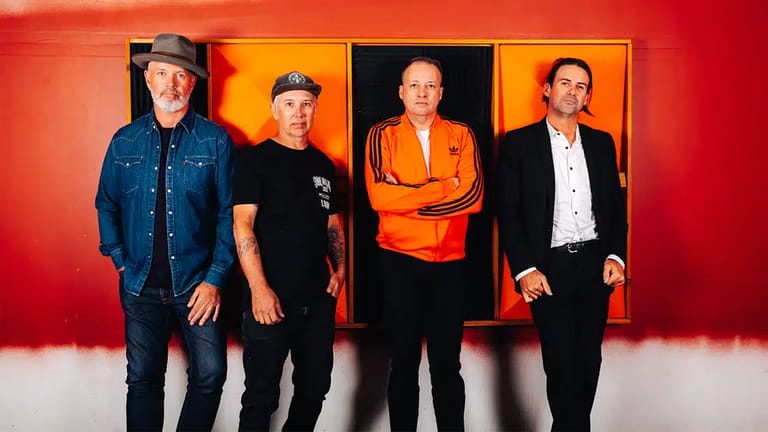 Four men stand against a vibrant red and orange gradient backdrop, wearing contrasting outfits from denim to formal wear.