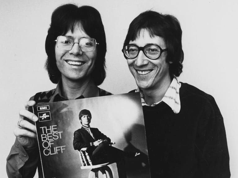 Two men with dark hair and glasses holding "The Best of Cliff" album cover, smiling at camera in black and white portrait.