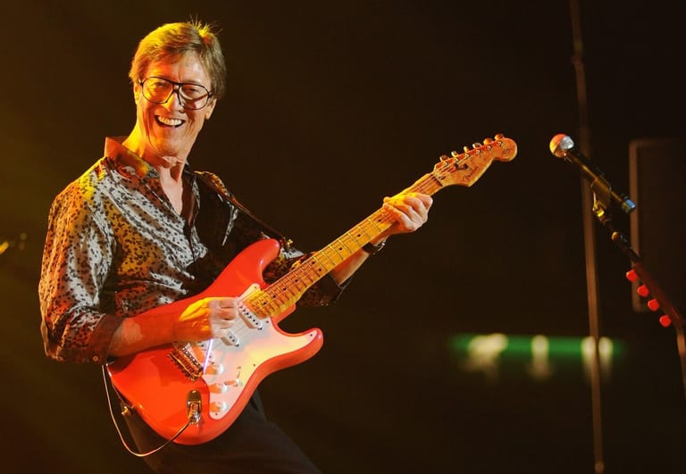 Musician with glasses playing a red electric guitar under warm stage lighting.