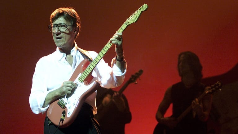 Guitarist in white shirt and glasses performs on stage against red lighting with bassist visible in shadow.