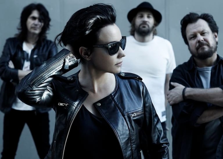 Four musicians posed against a grey background, with the front figure wearing sunglasses and a black leather jacket.