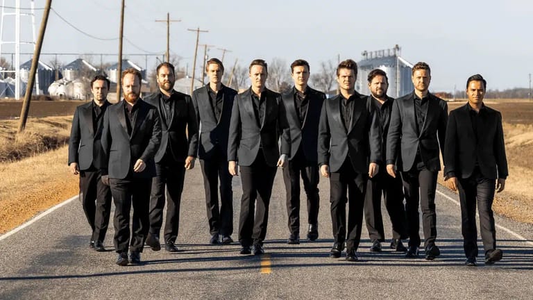 Ten men in dark suits walk down a rural road lined with power lines.
