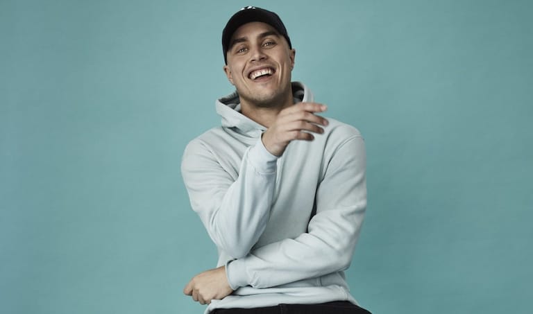 Man in light grey hoodie smiling against turquoise background.