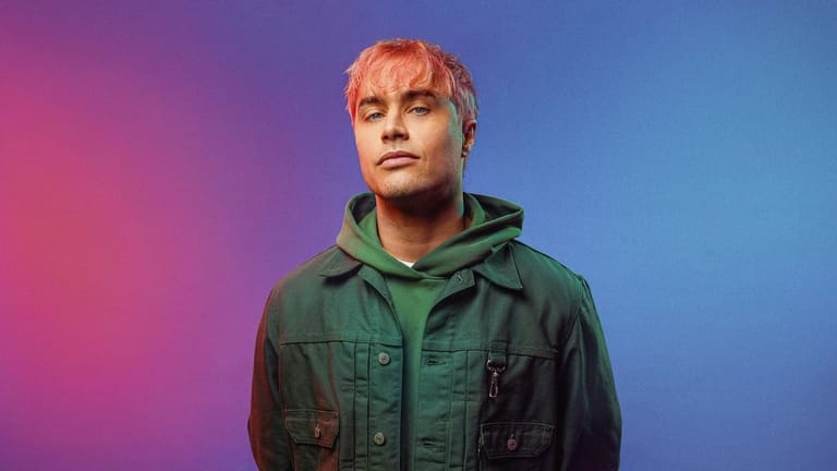 Young man with blonde hair wearing a dark green jacket against a gradient pink and blue background.