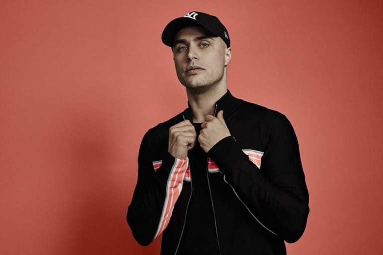 Man in black jacket and cap against coral pink background, adjusting striped scarf at neckline.