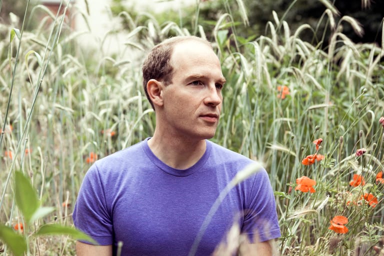 Man in purple shirt standing among tall grasses and orange wildflowers, gazing to the side.