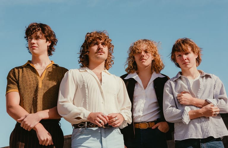 Four musicians with curly hair stand against a blue sky in casual vintage-style clothing.