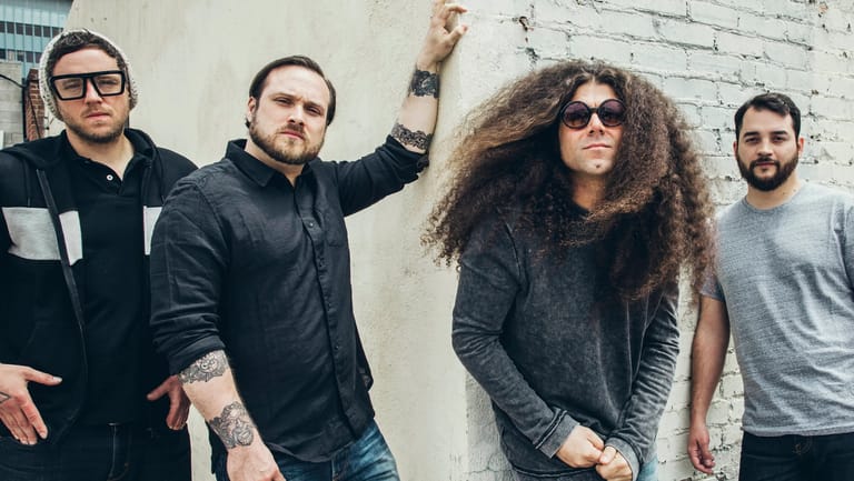 Four band members pose against a white brick wall, with one wearing sunglasses and curly hair in the centre.