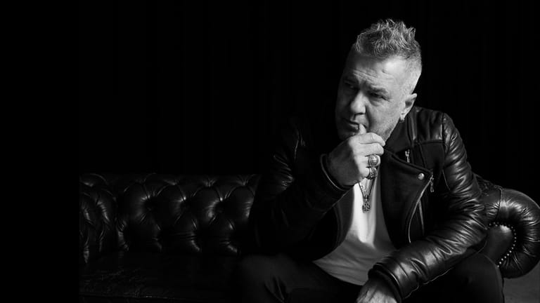 Man in black leather jacket seated on a sofa, hand to chin in contemplative pose, against dark background.