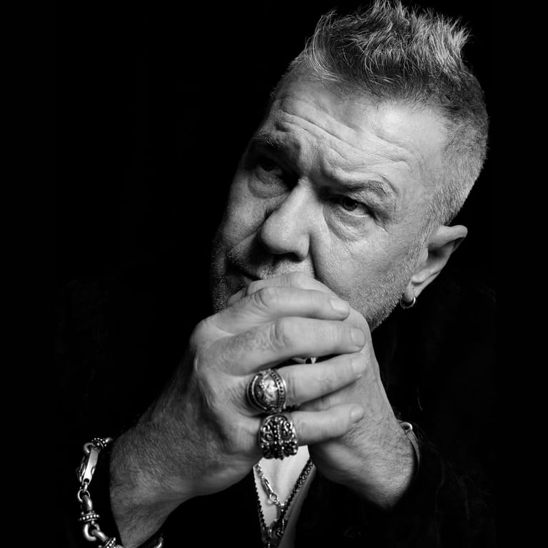 Middle-aged man in contemplative pose with hand to face against black background, wearing rings and bracelets.