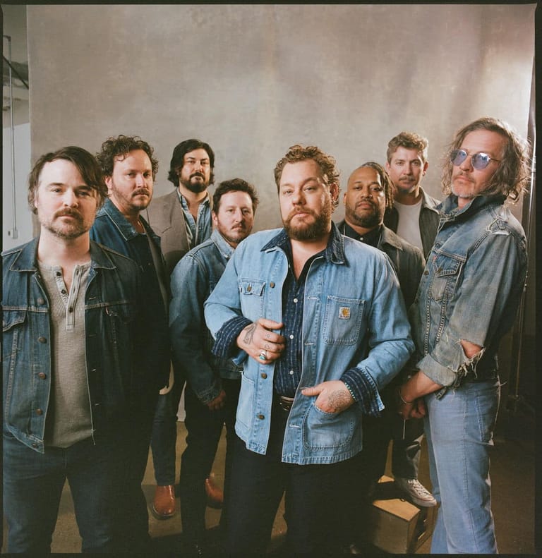 Eight men in denim and casual wear stand together against a neutral backdrop in a band portrait.