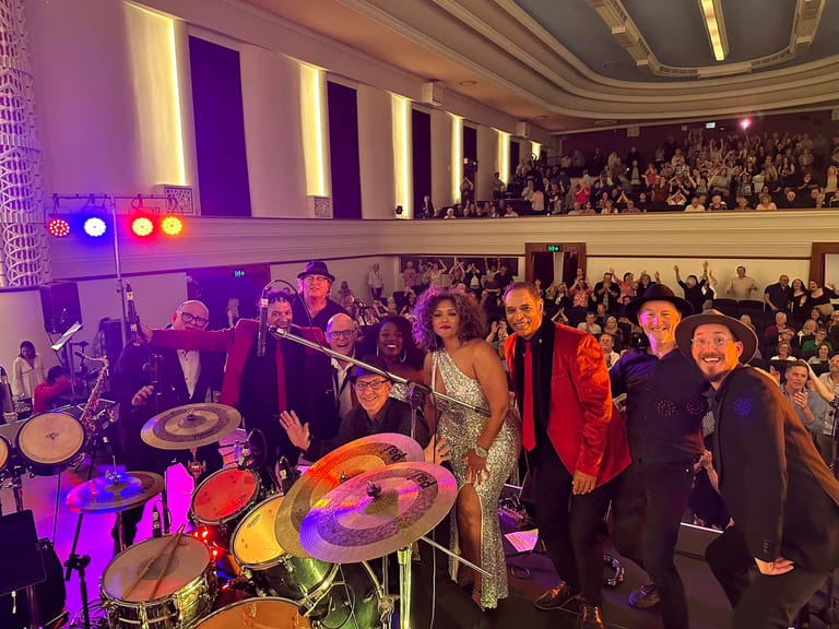 Live band performs on stage in ornate theatre with colourful lighting and packed audience in balcony.