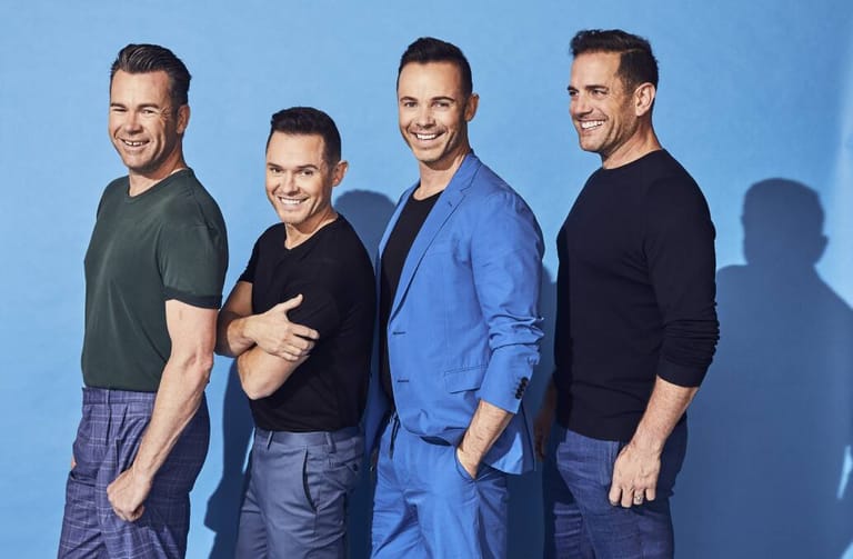 Four men in casual and smart-casual clothing pose together against a bright blue background.