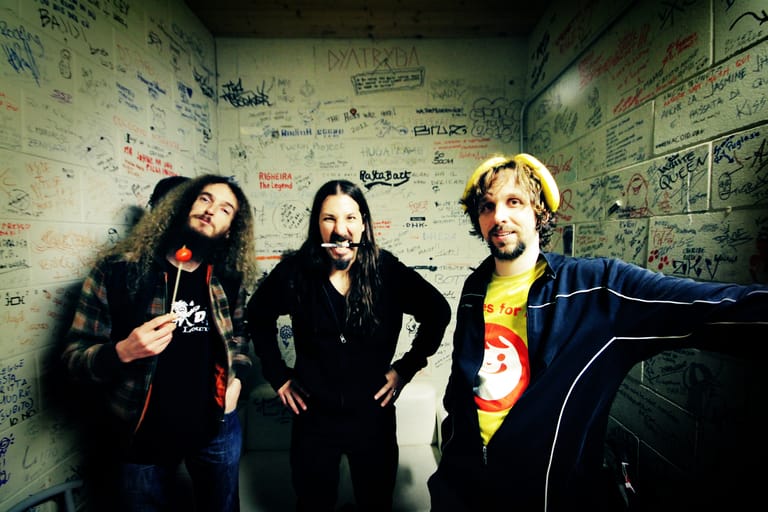 Three musicians with long hair stand against a wall covered in graffiti and band posters.