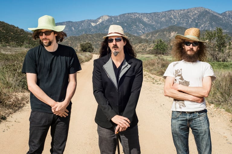 Three musicians in wide-brimmed hats stand on a dusty road with mountains in the background.