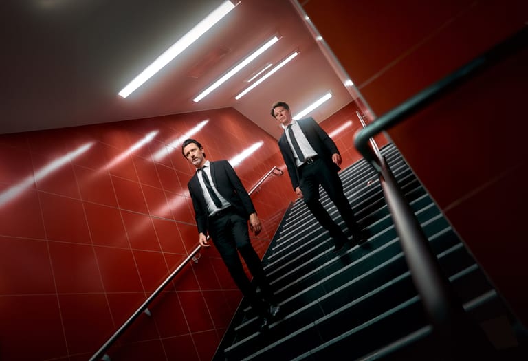 Two figures in dark suits ascending a staircase beneath geometric red and white ceiling lights.