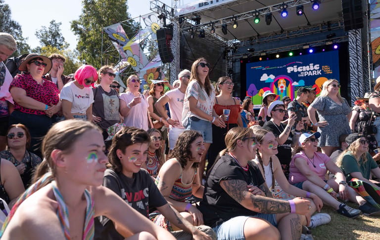 Outdoor festival crowd seated on grass facing a stage with performers and blue lighting under afternoon sun.