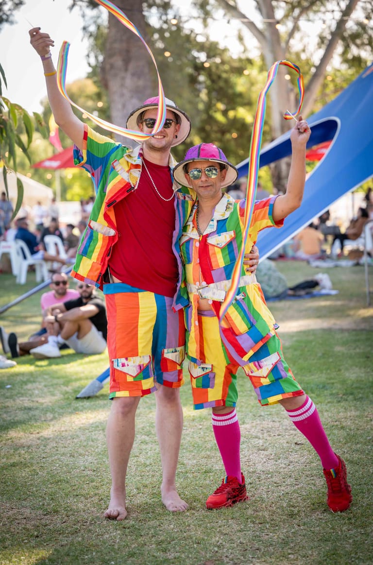 Two people in vibrant rainbow-striped costumes with oversized hats pose with ribbons at an outdoor park festival.