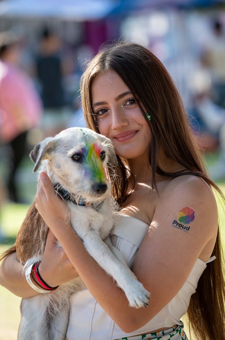 Woman holding a cream-coloured dog with rainbow paint on its face at an outdoor event.