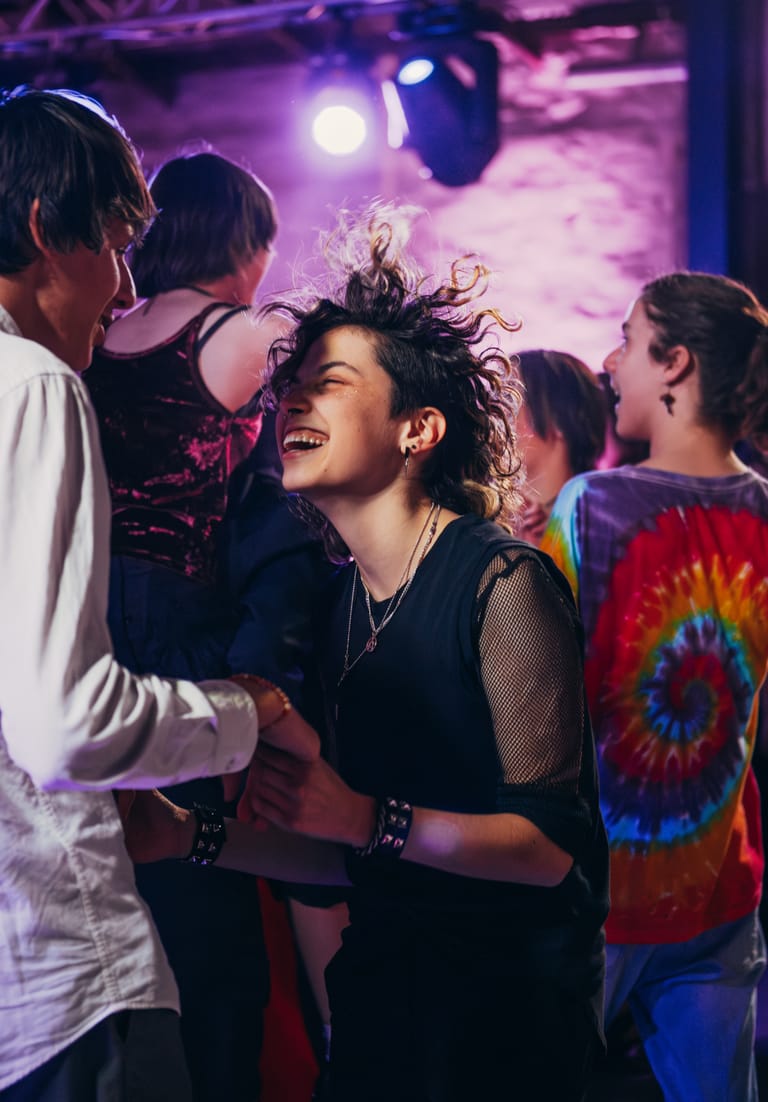 Smiling woman dancing amongst a crowd under purple stage lighting at a live music venue.