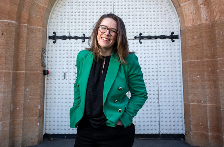 Woman in green blazer and black shirt standing in front of an arched brick doorway.