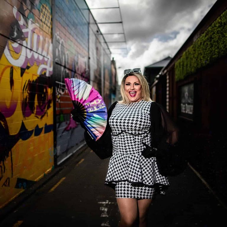 Woman in black-and-white houndstooth dress holding a rainbow fan stands in a laneway lined with vibrant street art and posters.