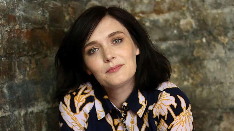 Woman with dark hair wearing a patterned black and gold jacket against a weathered brick wall.