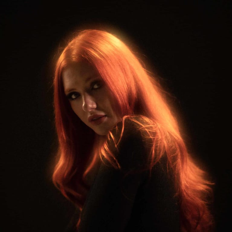 Person with long copper-red hair backlit by warm golden light against a black background.