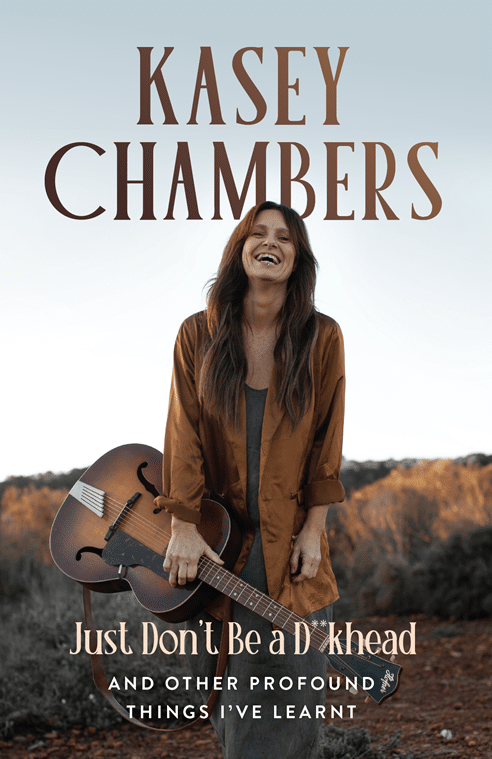Musician holding an acoustic guitar smiles outdoors in warm light, wearing a brown jacket against a golden landscape.