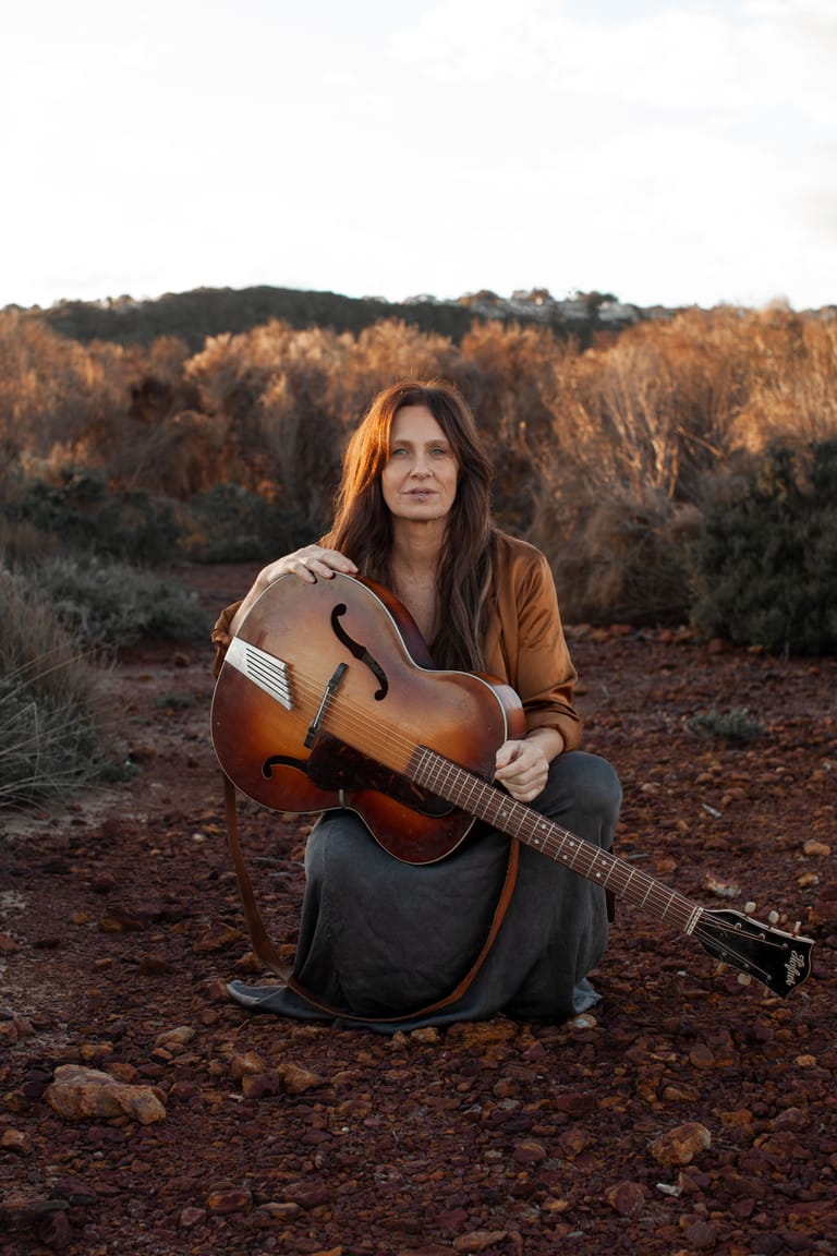 Musician seated outdoors holding an acoustic guitar amid golden-brown scrubland and dry grass.