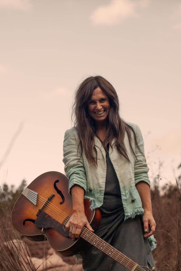 Musician holding an acoustic guitar outdoors, wearing a sage cardigan over dark clothing, smiling at camera against a muted sky.